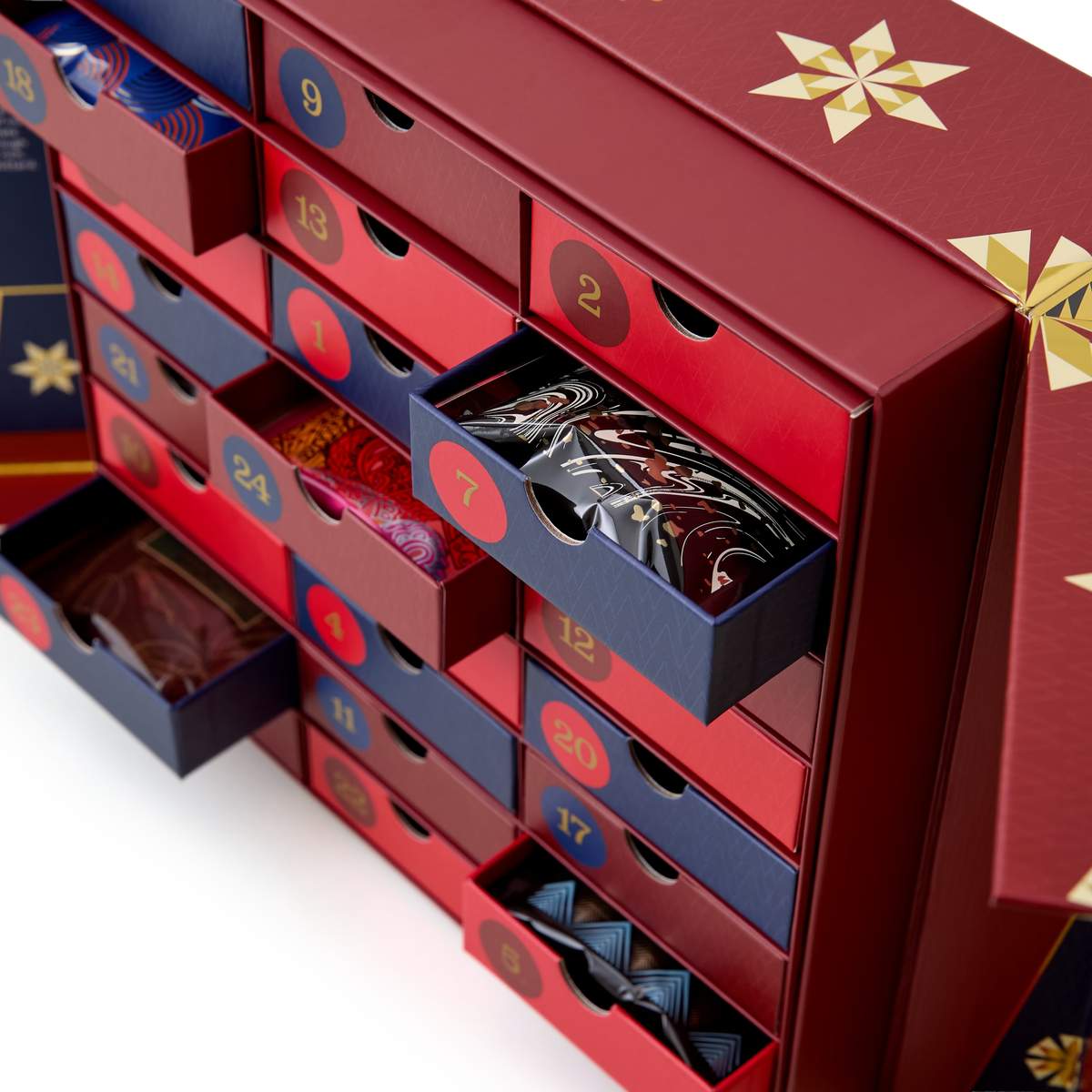 A close-up of the Hot Chocolate Advent Calendar showcases its red and blue numbered drawers, some opened to reveal an assortment of patterned chocolates, including festive flavors like carrot cake hot chocolate and white hot chocolate.