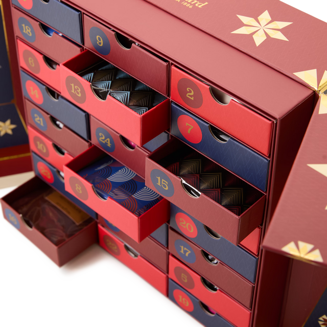 A close-up of the Hot Cocoa Advent Calendar in red and blue, featuring numbered drawers—some partially open to reveal patterned surprises inside. Gold accents decorate the box, making it a festive holiday cocoa gift.