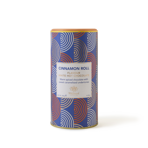 A cylindrical tin of Cinnamon Roll White Hot Chocolate featuring a blue, red, and white geometric-patterned packaging. The label describes this white chocolate drink as warmly spiced with sweet caramelised undertones, offering a delicious twist on classic cinnamon hot chocolate.