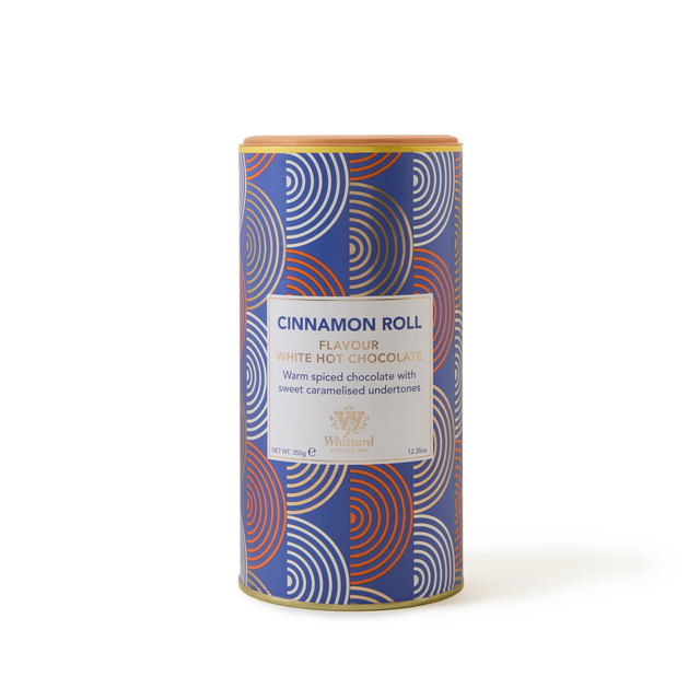 A cylindrical tin of Cinnamon Roll White Hot Chocolate featuring a blue, red, and white geometric-patterned packaging. The label describes this white chocolate drink as warmly spiced with sweet caramelised undertones, offering a delicious twist on classic cinnamon hot chocolate.