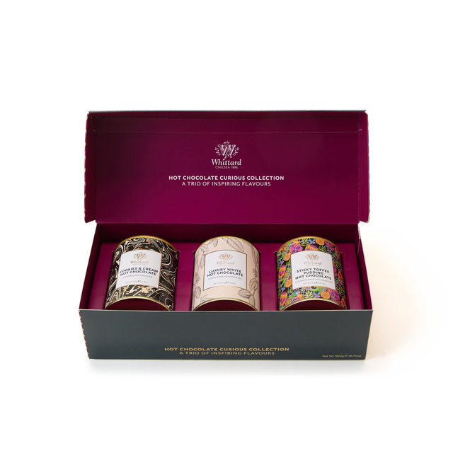 The Hot Chocolate Curious Collection features three tins of Whittard hot chocolate—including luxury white hot chocolate and cookies & cream hot chocolate—each in distinctively decorated containers, all arranged neatly inside an open magenta and black rectangular box on a white background.