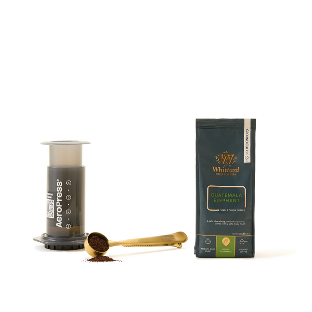 An AeroPress Coffee Starter Kit, a gold coffee scoop with ground coffee, and a bag of Whittard Guatemala Elephant ground coffee are arranged on a white background—perfect essentials for anyone passionate about exceptional coffee brewing.