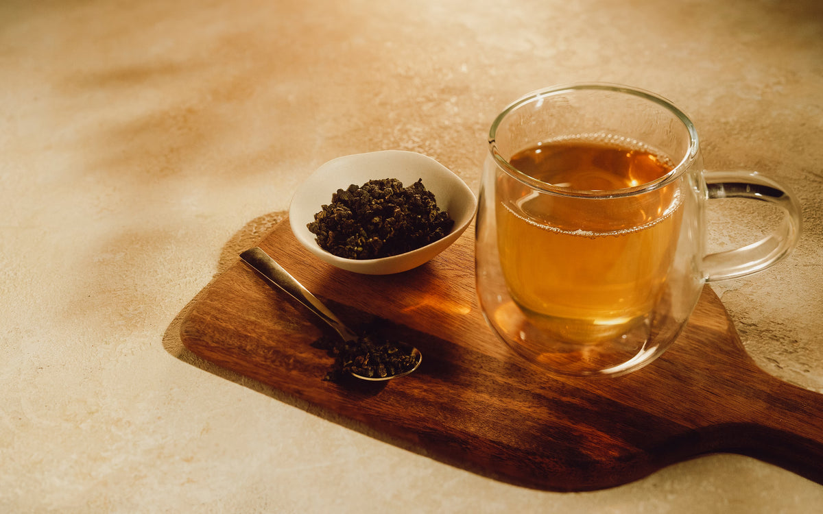A glass mug of tea, a small bowl of dry tea leaves, and a spoon with leaves on a wooden board.