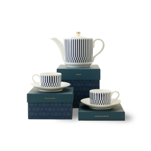 The Chelsea Collection Sloane Tea Set is displayed on blue boxes with gold lettering and understated patterns. The set includes a white teapot and two teacups, each adorned with blue geometric designs and resting on matching saucers. Subtle 22ct gold detailing accentuates each piece, while the plain white background highlights the elegance of British ceramic craftsmanship.