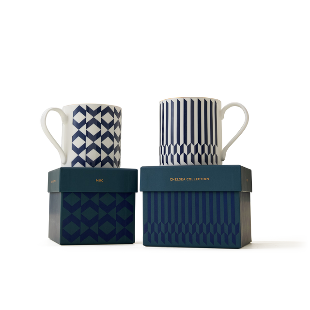 Two white mugs adorned with blue geometric patterns and elegant 22ct gold detailing sit atop matching blue boxes labeled “MUG” and “CHELSEA COLLECTION,” set against a plain white backdrop, showcasing the refined style of The Chelsea Collection Mug Duo and celebrating the excellence of British craftsmanship.