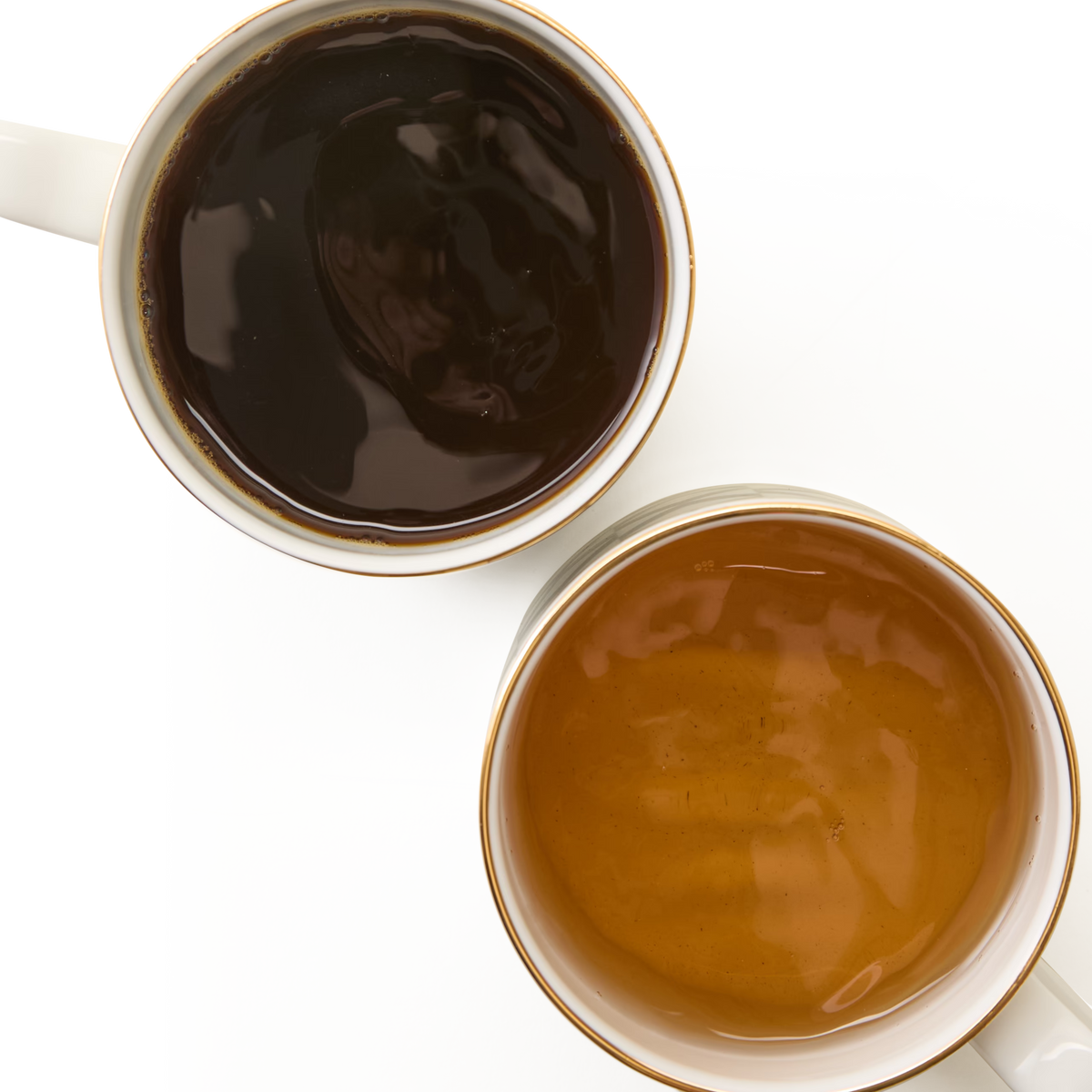 The Chelsea Collection Mug Duo, celebrated for its British craftsmanship and exquisite 22ct gold detailing, is displayed on a white background—one mug brimming with rich black coffee and the other with creamy light brown coffee—placed diagonally and gently overlapping.