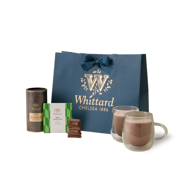 Introducing The Chocolate Indulgence Set: a luxurious hot chocolate gift set that includes a blue Whittard Chelsea 1886 bag, a tin of single origin Colombian milk chocolate hot chocolate, a box of assorted chocolates, several irresistible chocolate pieces, and two elegant glass mugs brimming with rich, velvety hot chocolate.