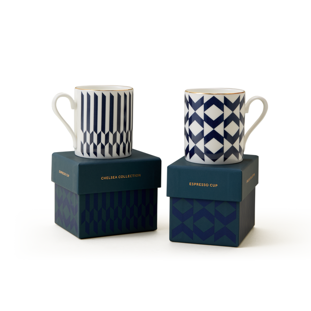 Two white espresso cups with blue geometric patterns sit atop blue boxes labeled “The Chelsea Collection Espresso Cup Duo.” Showcasing exquisite British craftsmanship, the boxes also feature dark blue geometric designs, all set against a plain white background.