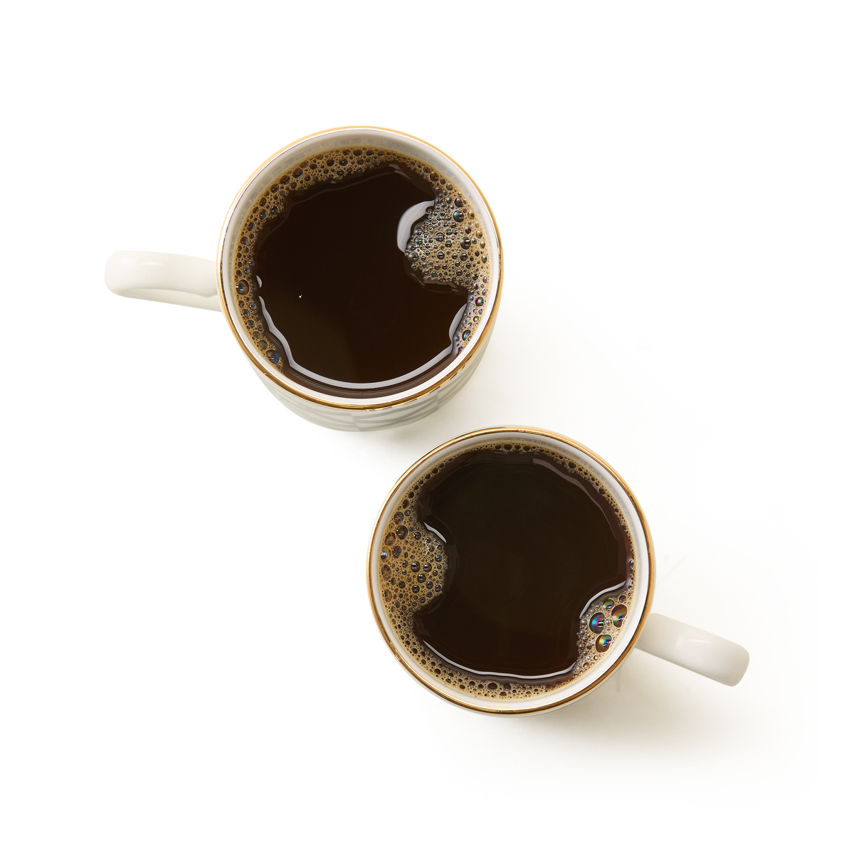 Showcasing British craftsmanship, the Chelsea Collection Espresso Cup Duo features two white ceramic mugs filled with black coffee, viewed from above and placed on a white surface. One larger mug is positioned above the smaller mug—reminiscent of elegant espresso cups—with subtle reflections and bubbles in the coffee completing this refined scene.