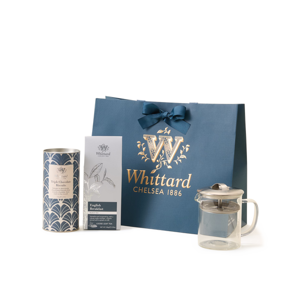 The Luxury Tea Time Gift Set is displayed on a white background, featuring a blue Whittard Chelsea 1886 gift bag, a tea gift box, a tin of loose leaf tea, and an elegant glass teapot arranged in front.
