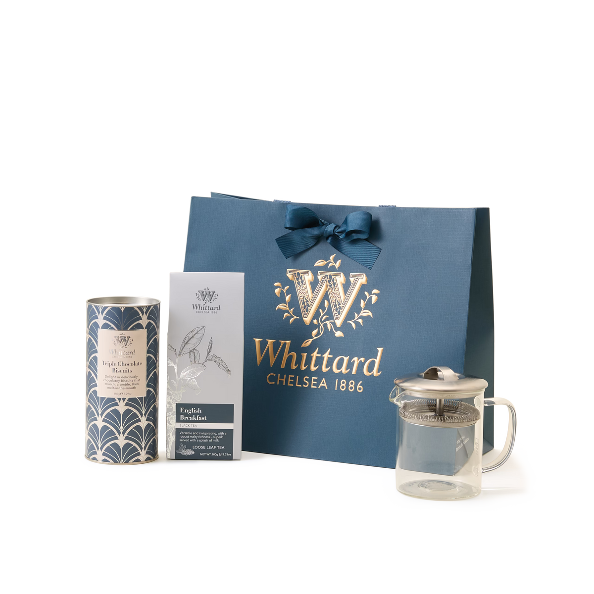 The Luxury Tea Time Gift Set is displayed on a white background, featuring a blue Whittard Chelsea 1886 gift bag, a tea gift box, a tin of loose leaf tea, and an elegant glass teapot arranged in front.
