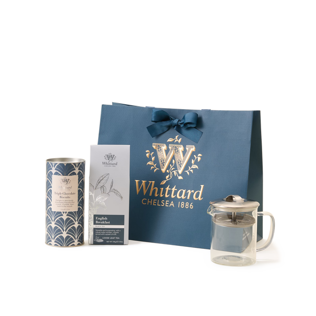 The Luxury Tea Time Gift Set is displayed on a white background, featuring a blue Whittard Chelsea 1886 gift bag, a tea gift box, a tin of loose leaf tea, and an elegant glass teapot arranged in front.