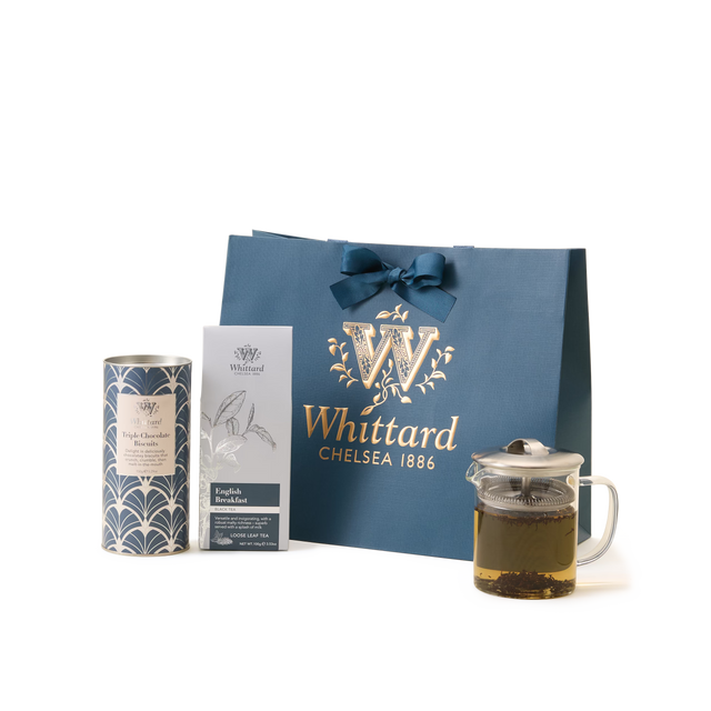 A Whittard gift set with a blue and gold bag, a tin of hot chocolate, a box of English Breakfast tea, and a glass teapot with brewed tea inside, all on a white background.