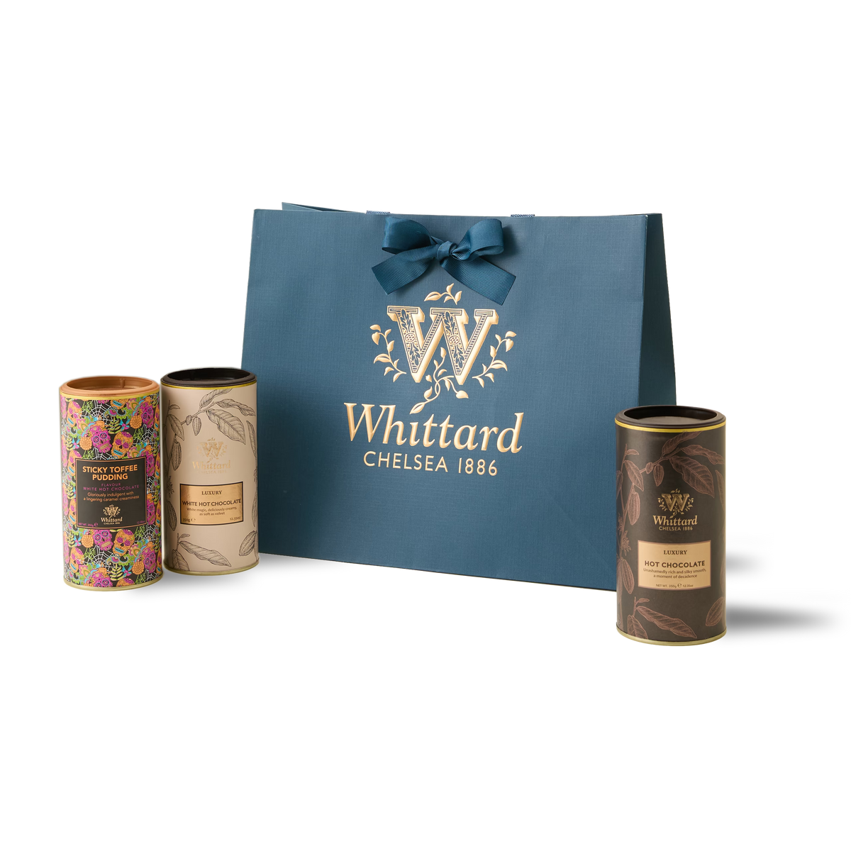 The Signature Hot Chocolate Trio features three cylindrical tins—Sticky Toffee Pudding Hot Chocolate, Luxury White Hot Chocolate, and Luxury Hot Chocolate—each adorned with colorful and gold designs, all presented behind a blue Whittard gift bag topped with a bow.