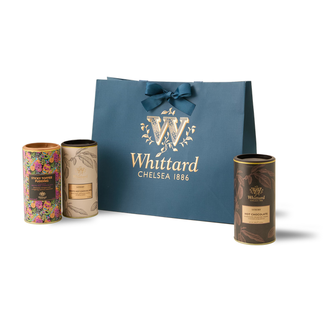 The Signature Hot Chocolate Trio features three cylindrical tins—Sticky Toffee Pudding Hot Chocolate, Luxury White Hot Chocolate, and Luxury Hot Chocolate—each adorned with colorful and gold designs, all presented behind a blue Whittard gift bag topped with a bow.
