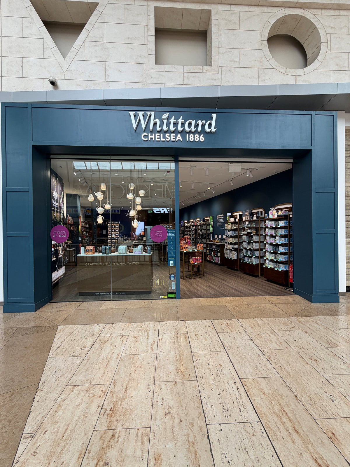 Whittard Chelsea 1886 store with a dark blue entrance, glass doors, and shelves of tea products inside. The interior is well-lit with hanging lights and displays.