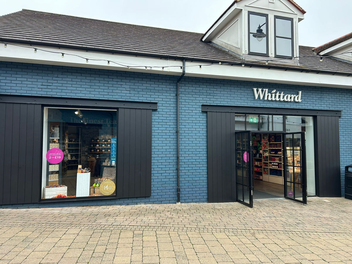 Exterior of a Whittard store with blue brick walls, large windows displaying products, and double doors open to the shop interior. Paved sidewalk is visible in the foreground.