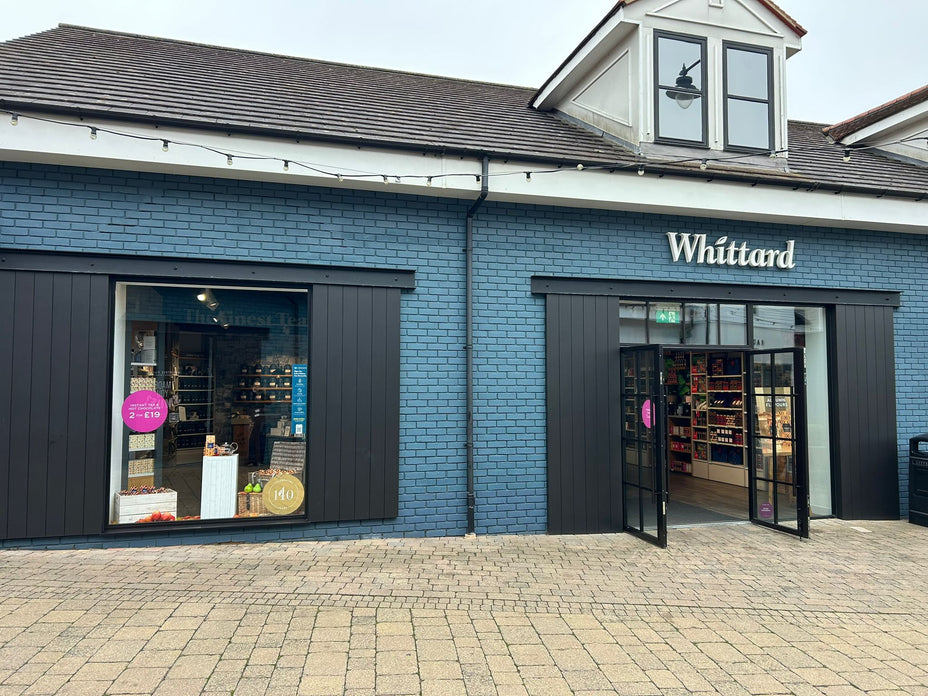 Exterior of a Whittard store with blue brick walls, large windows displaying products, and double doors open to the shop interior. Paved sidewalk is visible in the foreground.
