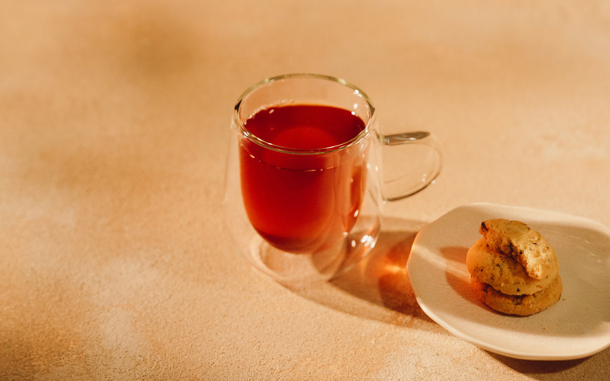 A glass mug of red tea next to a small plate with three stacked cookies.