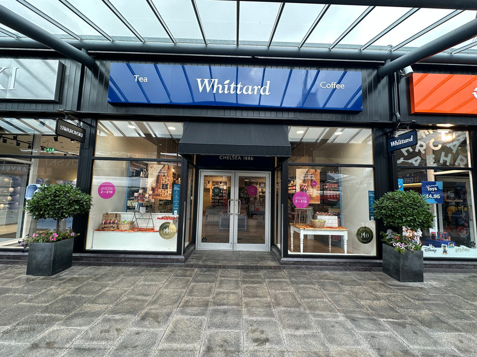 A Whittard tea and coffee shop with glass doors, large blue signage, and potted plants outside. The shop interior displays shelves of products and promotional signs in the windows.
