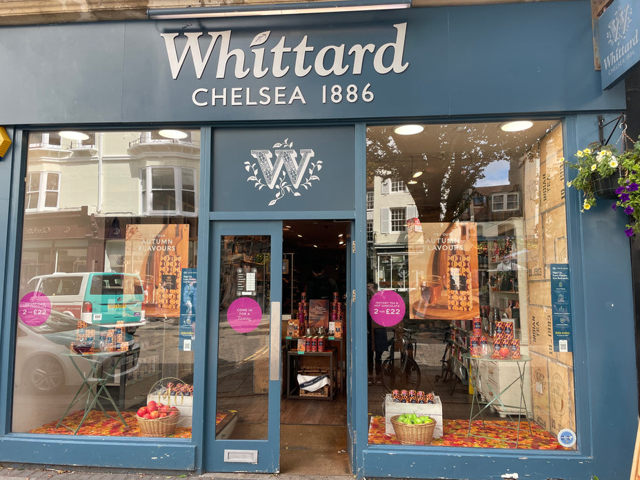 Whittard Chelsea 1886 storefront with glass windows, autumn-themed displays, posters advertising offers, baskets of apples, and various tea products visible inside. The shop is on a street with cars and buildings reflected in the glass.