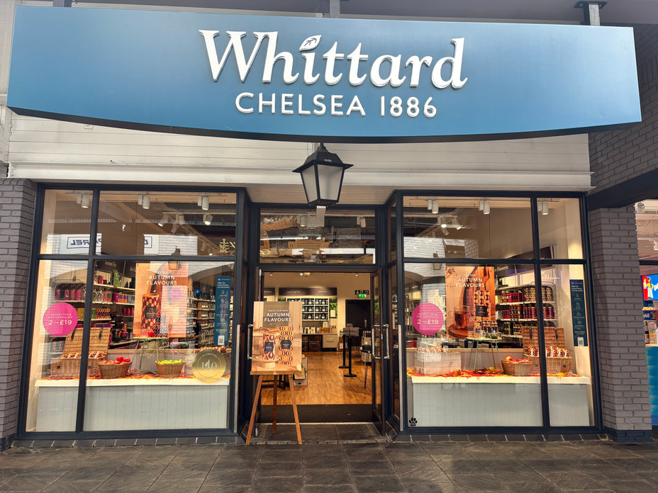 Storefront of Whittard Chelsea 1886 with glass windows, displays of tea products, promotional signs, and shelves with colorful tins inside. Blue sign with white lettering is above the entrance.