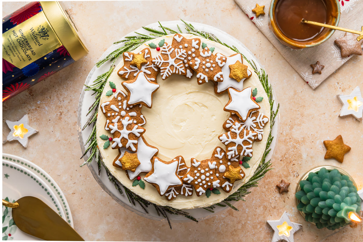 A round cake with white icing is decorated with gingerbread cookies shaped like stars and snowflakes. Rosemary sprigs circle the cake. A cup of caramel, candle, and festive tin are nearby.