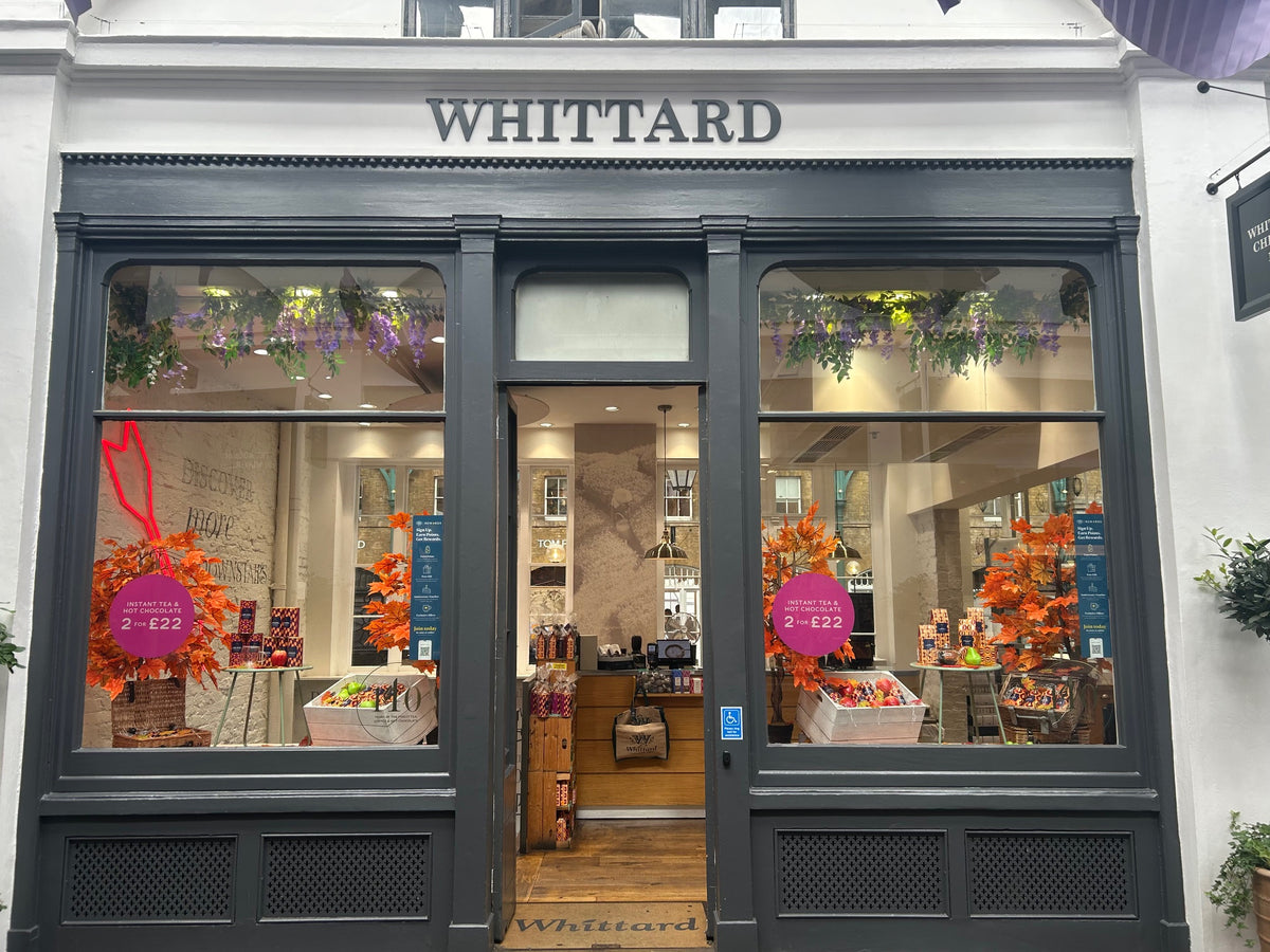 Shopfront of Whittard with large windows displaying autumn-themed decorations, baskets of products, and signs reading 2 for £22. Greenery hangs above the entrance and purple flowers decorate the top of the windows.
