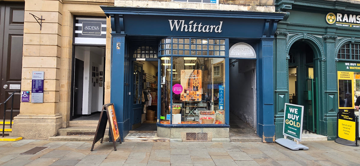 A blue storefront labeled Whittard displays tea and products in the window. Signs and advertisements are outside on the pavement, and the shop sits between other businesses on a stone street.