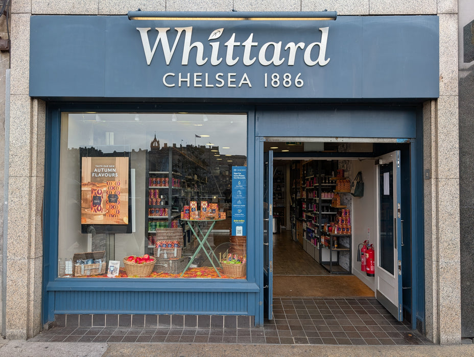 Storefront of Whittard Chelsea 1886 with large windows displaying products and a stand advertising autumn flavors. The entrance door is open, revealing shelves stocked with goods inside.