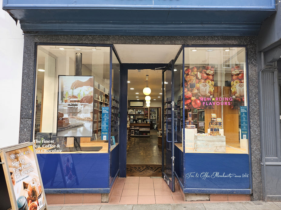 A storefront window display for Tea & The Market, featuring blue-framed glass doors and windows, a tiled entryway, and interior shelving visible behind. A sample board with baked goods stands on the left sidewalk.