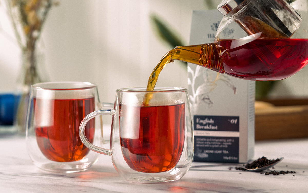 A glass teapot pours amber tea into a glass mug beside another filled mug. A box of English Breakfast tea and loose tea leaves sit on a white countertop in soft natural light.