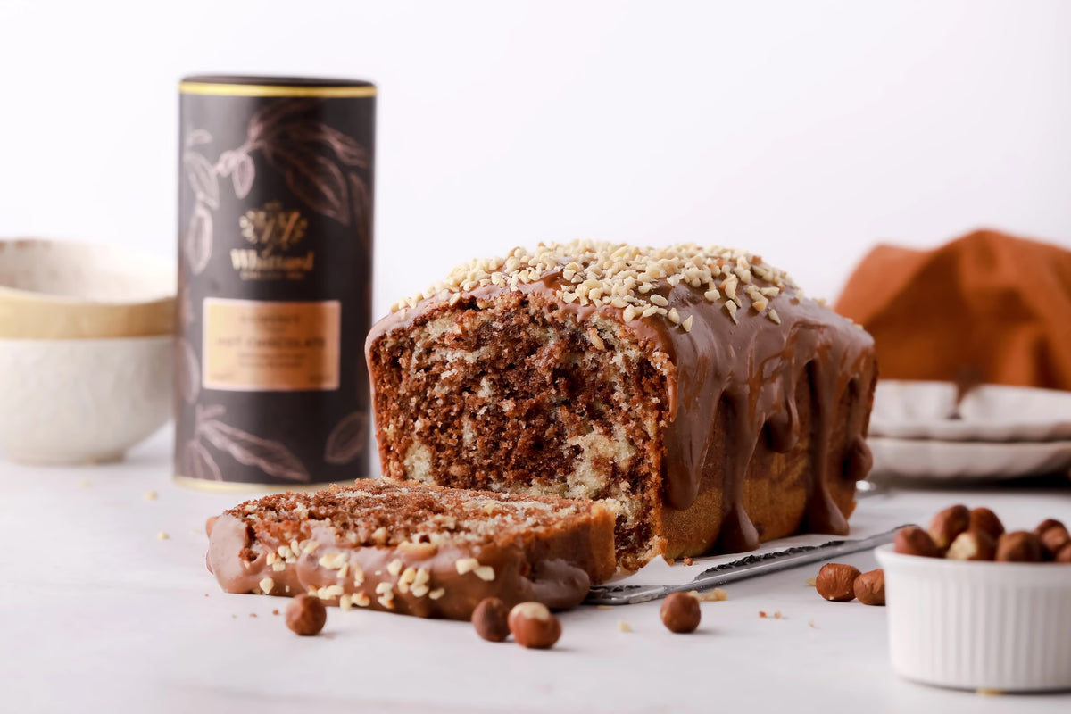 A marble loaf cake with chocolate glaze and chopped nuts on top, with one slice cut. Hazelnuts are scattered nearby, and a black canister and bowls are in the background.