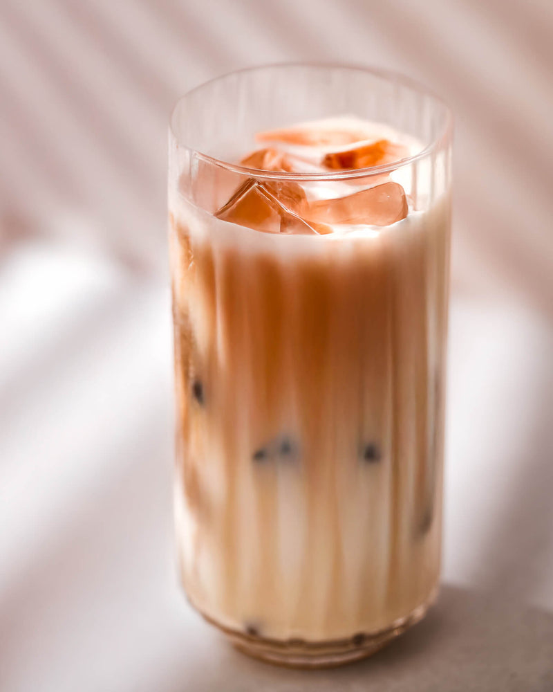 A tall glass filled with iced milk tea, showing layers of creamy milk and tea, with several ice cubes floating on top. The glass stands on a light surface with soft, striped shadows in the background.