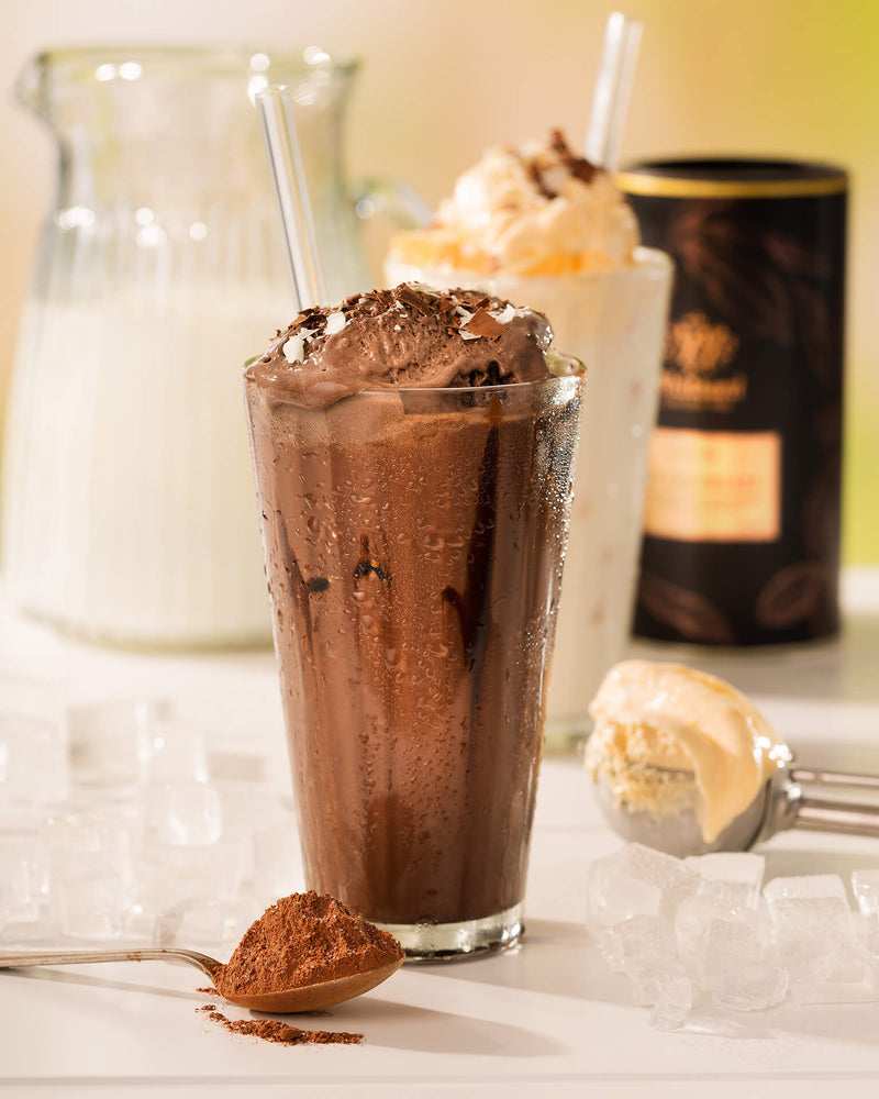 Iced chocolate milkshake with chocolate shavings in a glass, surrounded by ice cubes, a spoonful of cocoa powder, a scoop of ice cream, and a vanilla milkshake in the background.