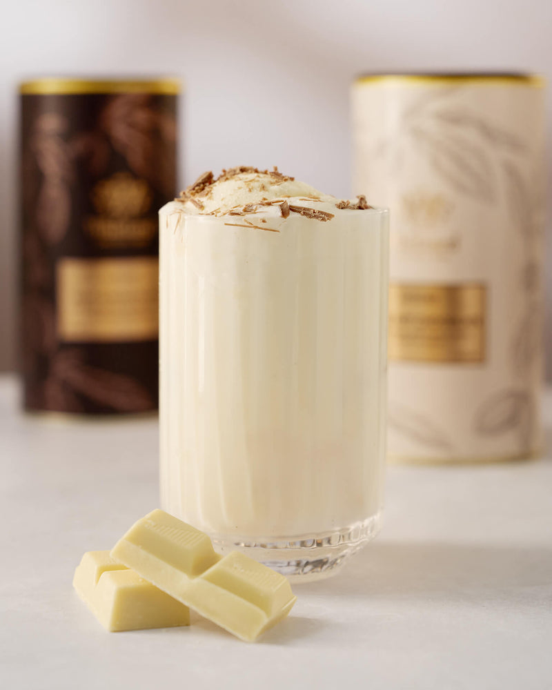 A glass of creamy white beverage topped with chocolate shavings sits beside pieces of white chocolate, with two blurred canisters in the background.