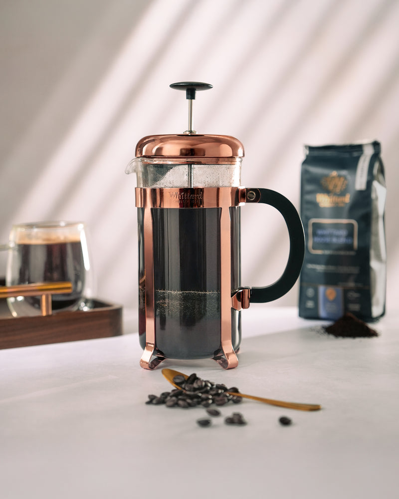 A copper French press with coffee sits on a white surface. Coffee beans and a spoon are in front, with a coffee bag, glass of coffee, and shadows from window blinds in the background.