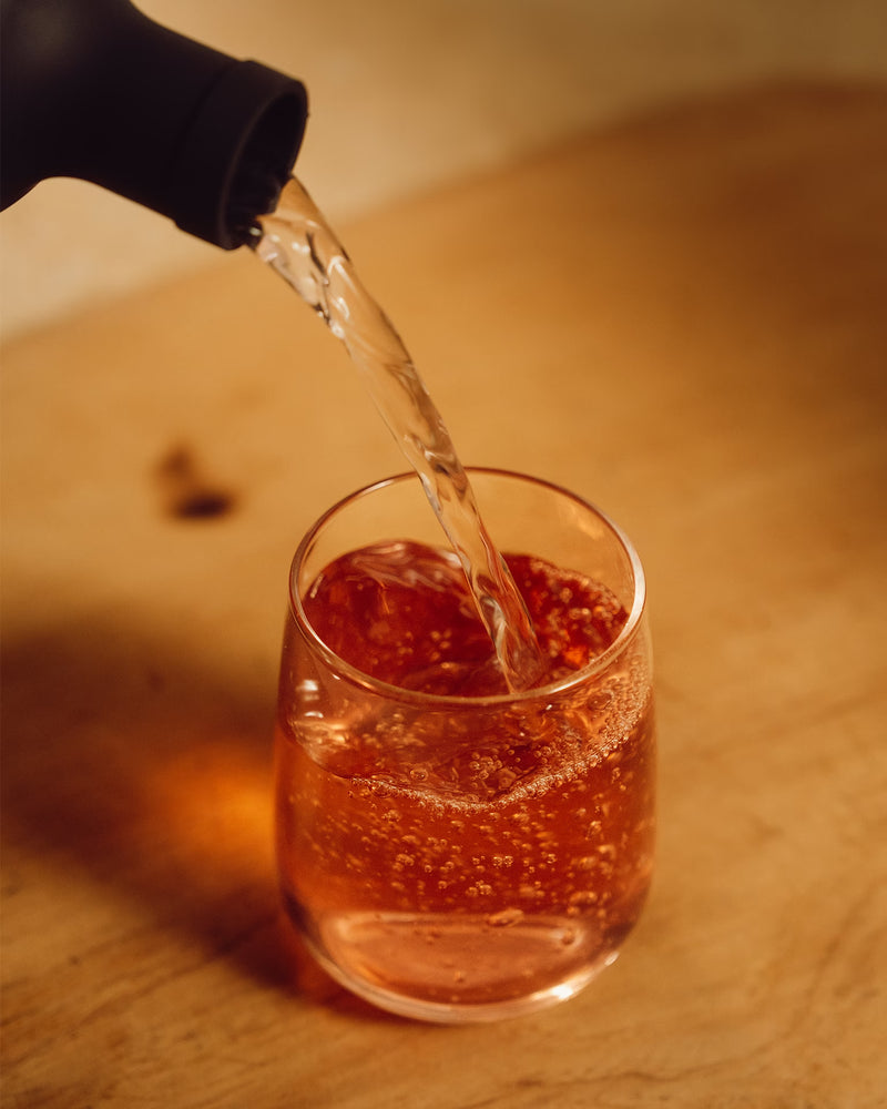 A clear liquid is poured from a bottle into a glass containing an amber-colored, carbonated beverage.