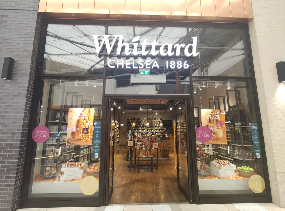 Storefront of Whittard Chelsea 1886 with glass doors and windows, displaying shelves of tea, coffee, and gifts inside. Promotional signs on the glass advertise offers. The entrance is framed by dark brick and cream walls.