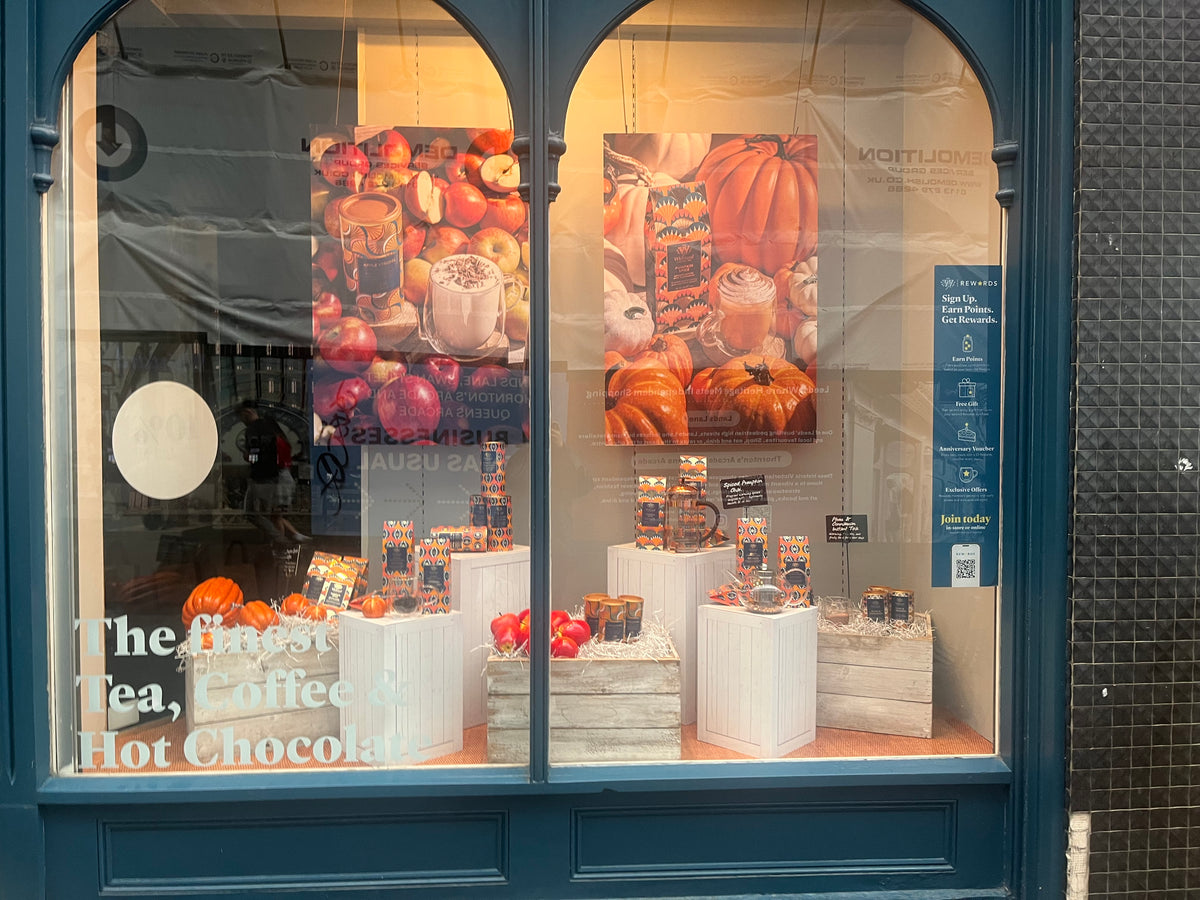 A storefront window display with blue trim showcasing packaged tea, coffee, and hot chocolate products arranged in white boxes and crates. Two autumn-themed posters with pumpkins and fruit hang behind the display, and large white text on the lower left reads 'The Best Tea, Coffee & Hot Chocolate'.