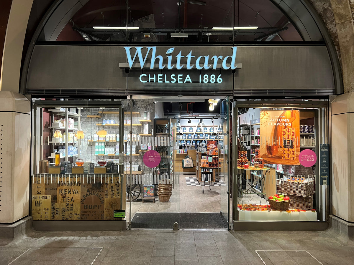 Storefront of Whittard Chelsea 1886, a tea and coffee shop, with glass doors, shelves of products inside, and autumn-themed displays featuring orange signage and baskets of apples near the entrance.
