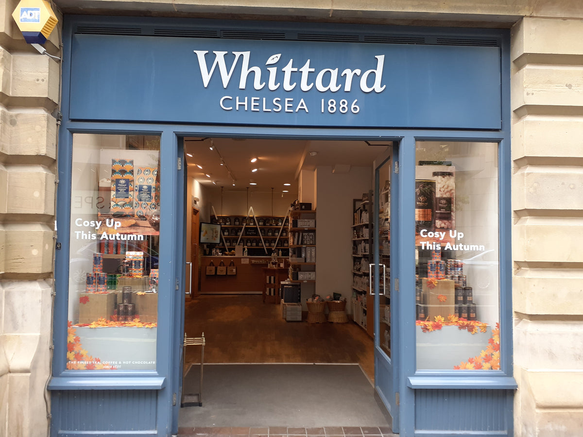 The storefront of Whittard Chelsea 1886, featuring a dark blue sign and door frame with white lettering, set against a beige stone facade. The open doorway reveals the warm interior of the shop with bookshelves and products.