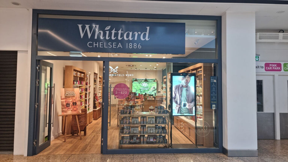 Whittard Chelsea 1886 store with glass front, shelves of tea and coffee products, and promotional signs inside. Display window shows shop interior and a large poster of a man holding a mug.