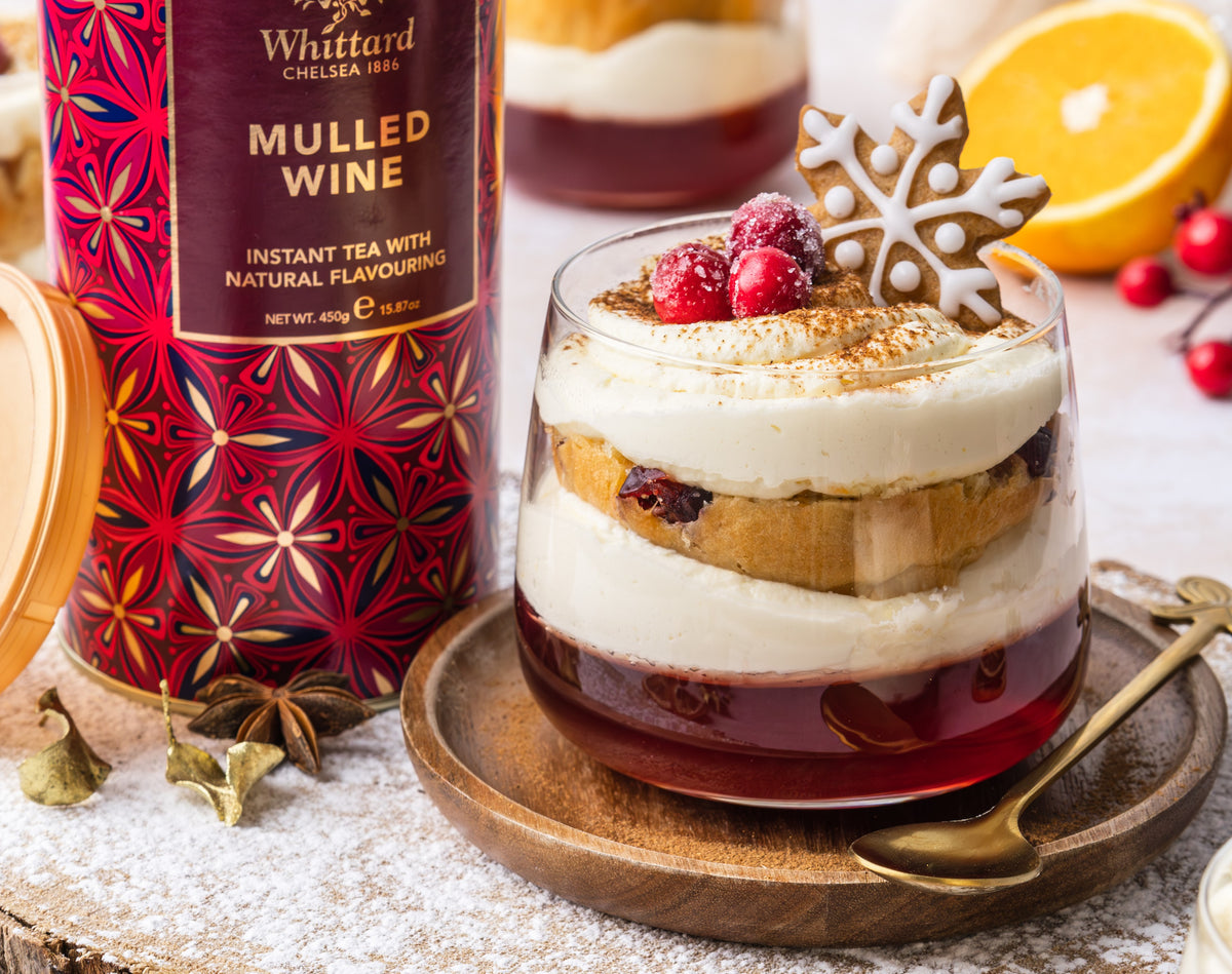 A glass dessert with cream, cake, red jelly, cranberries, and a snowflake cookie, next to a container of Whittard Mulled Wine instant tea. An orange half and festive spices are also on the table.