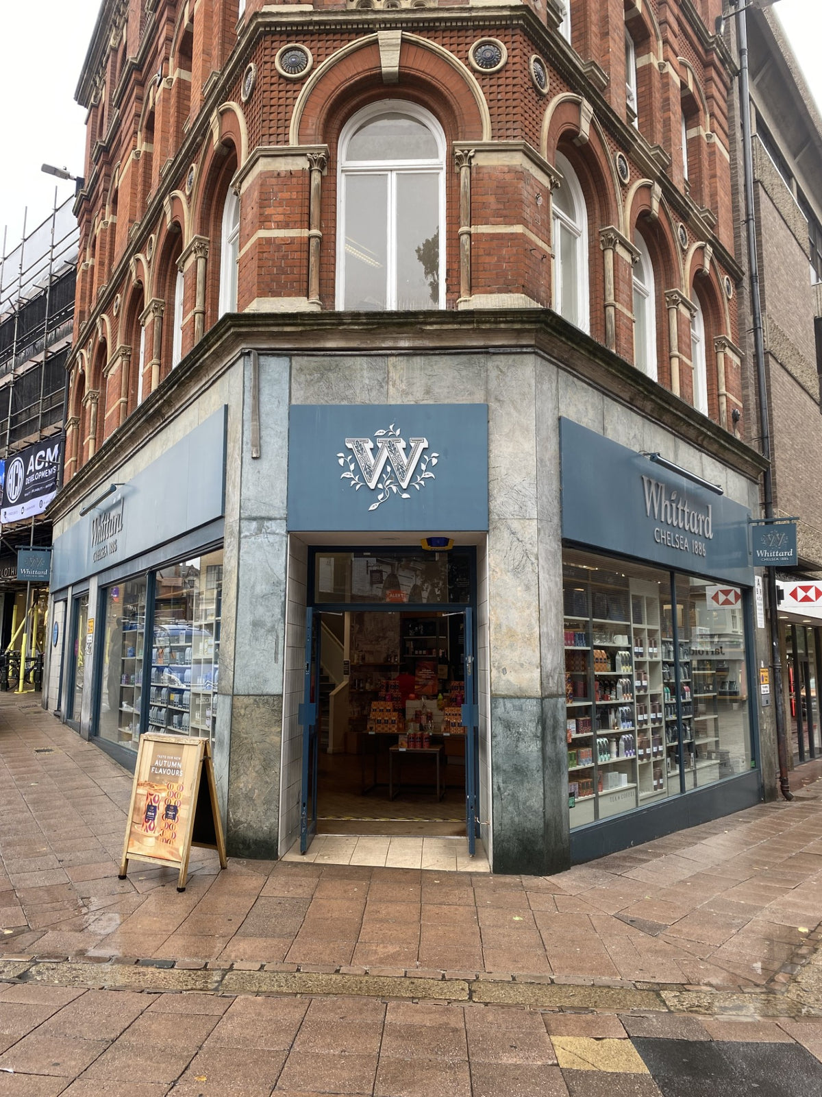A Whittard of Chelsea store on a street corner with large windows, a blue sign featuring a white W logo, and products visible inside. The surrounding pavement is wet from recent rain.