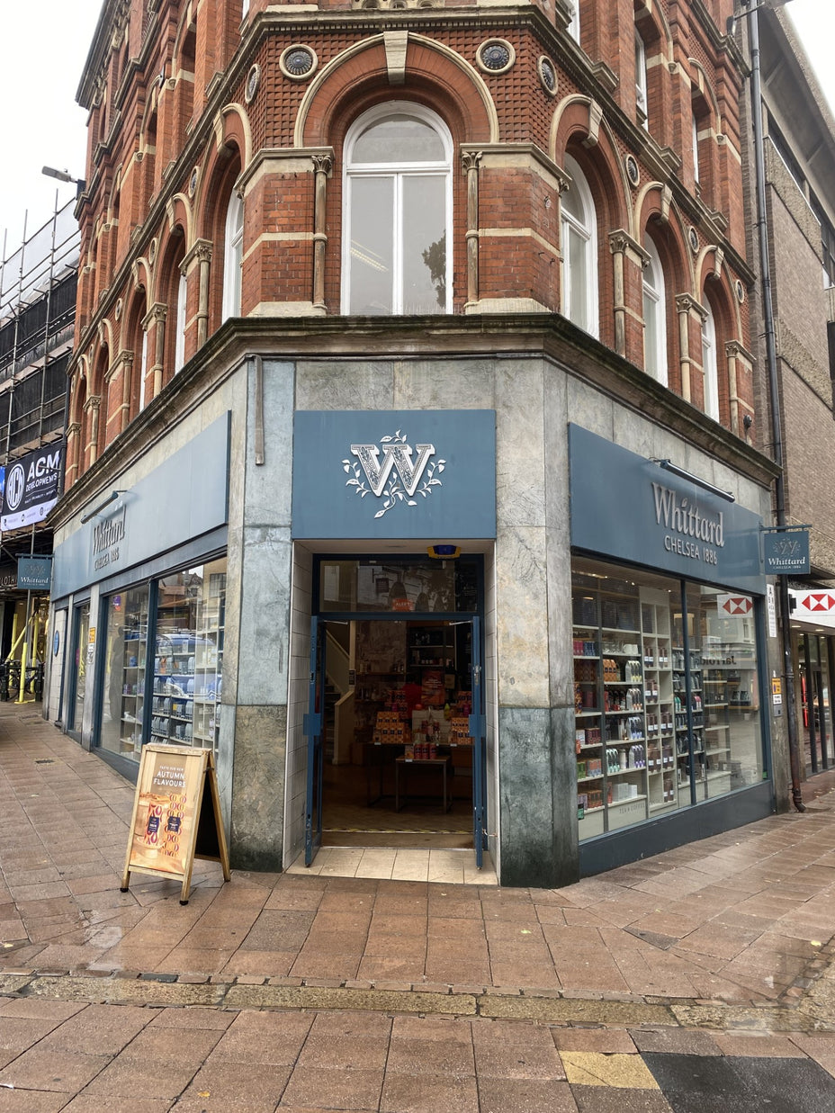 A Whittard of Chelsea store on a street corner with large windows, a blue sign featuring a white W logo, and products visible inside. The surrounding pavement is wet from recent rain.