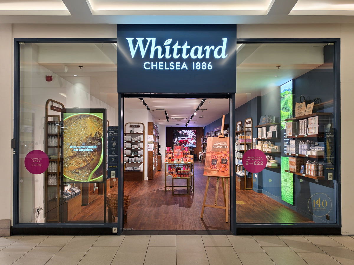 Whittard Chelsea 1886 storefront with glass doors, product displays, colorful packaging, and shelves of tea, coffee, and gifts visible inside a shopping mall setting.