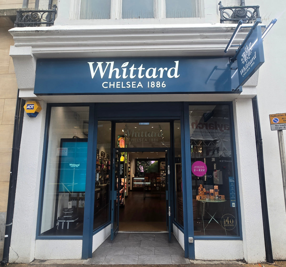 Street view of Whittard Chelsea 1886 shopfront with a blue sign above glass doors, displaying teas and products inside. A promotional poster and stack of boxes are visible through the window.