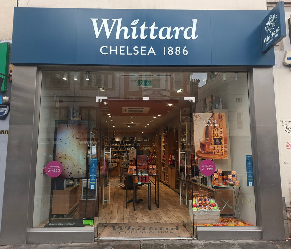 Storefront of Whittard Chelsea 1886 with glass doors, shelves of tea and related products inside, and promotional signs on the entrance. The shop has a blue sign with white text above the door.