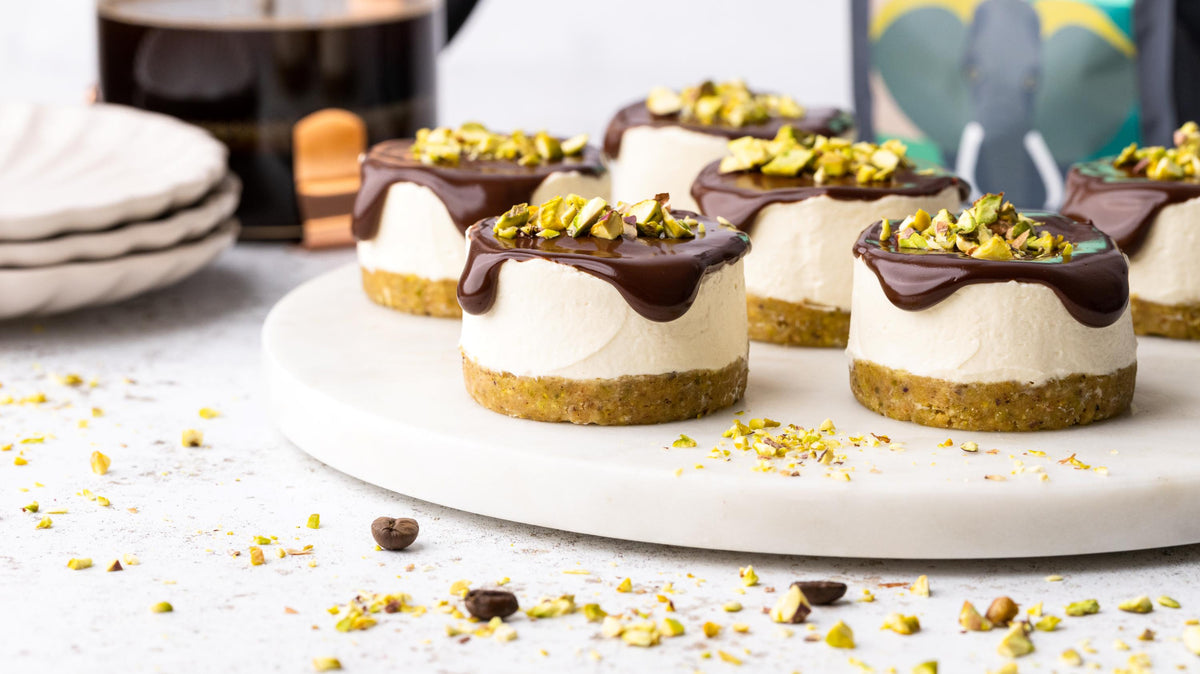 Mini cheesecakes with a nut crust, topped with chocolate ganache and chopped pistachios, arranged on a white platter. Scattered pistachios, plates, and a coffee press are in the background.